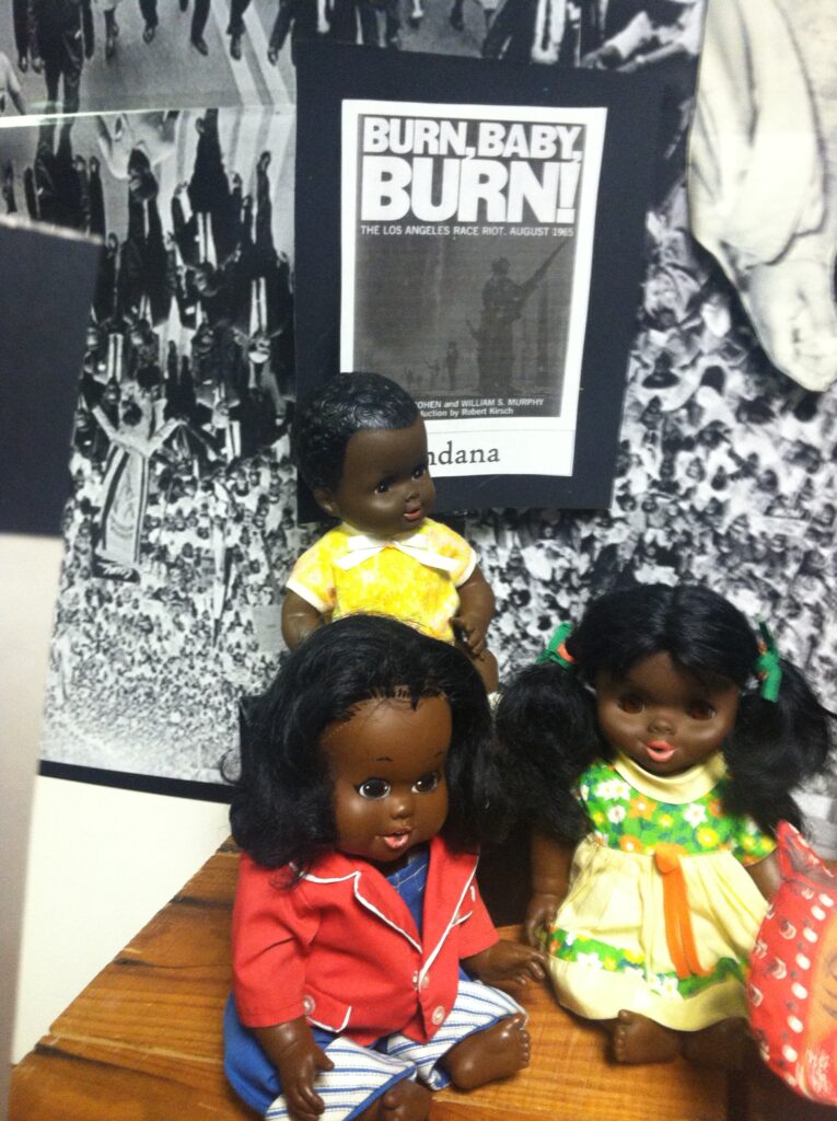 Three Black dolls in front of riot poster